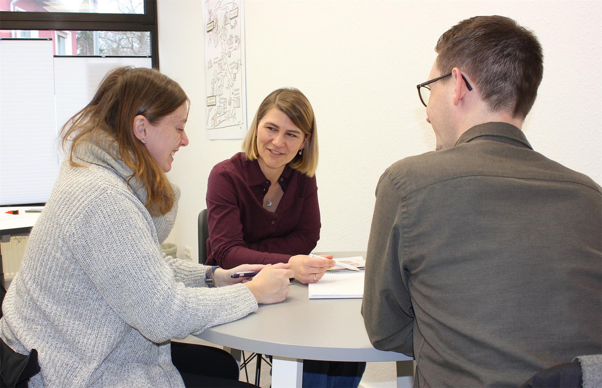 Schwangerschaftsberatung in einem hellen Büro mit drei Personen, die an einem runden Tisch ein persönliches Gespräch führen. Die Beraterin in der Mitte hält einen Stift in der Hand, während die beiden Ratsuchenden aufmerksam zuhören und Notizen machen.