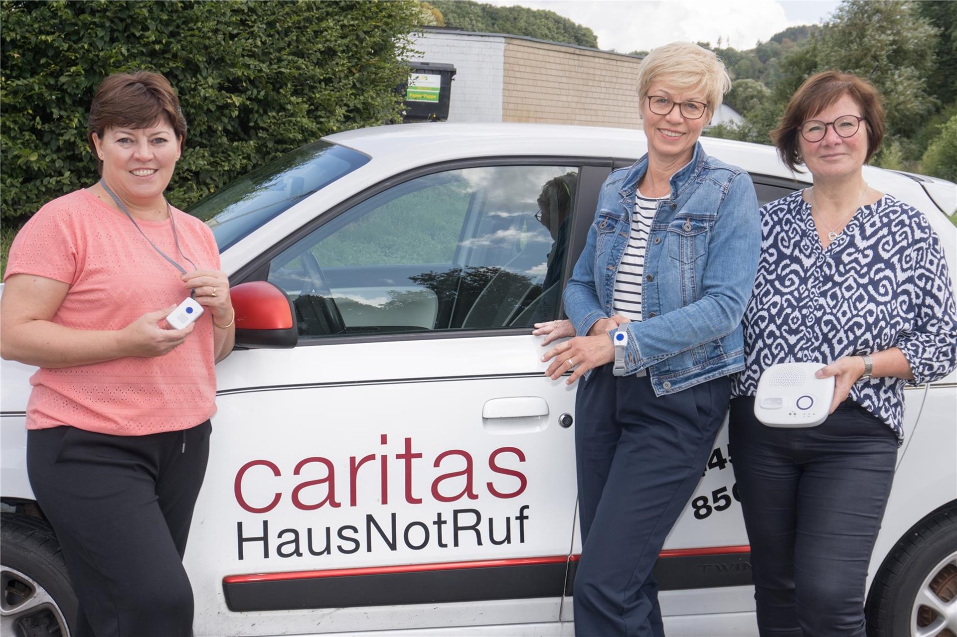 Die drei HausNotRuf-Mitarbeiterinnen Helena Fast (links), Brigitte Dovern (Mitte) und Marita Breuer (rechts) stehen vor ihrem Dienstwagen mit der Aufschrift "Caritas HausNotRuf". Sie halten verschiedene Teile des HausNotRuf-Systems in der Hand.