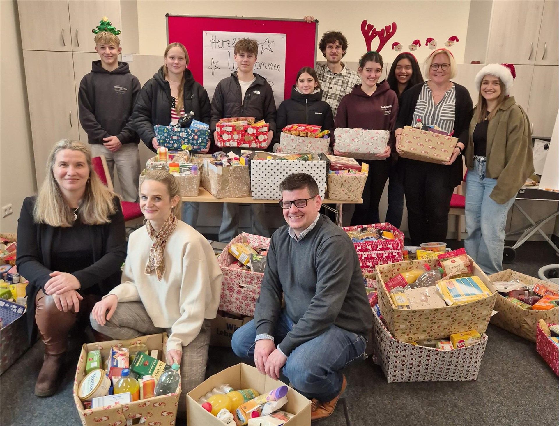 Schüler und Lehrkräfte der Clara-Fey-Schule sowie Mitarbeitende der Caritas stehen bzw. um die zahlreichen gespendete Lebensmittelpakete herum und lächeln in die Kamera. Teilweise haben sie Weihnachtsmützen und andere weihnachtliche Gegenstände auf. 