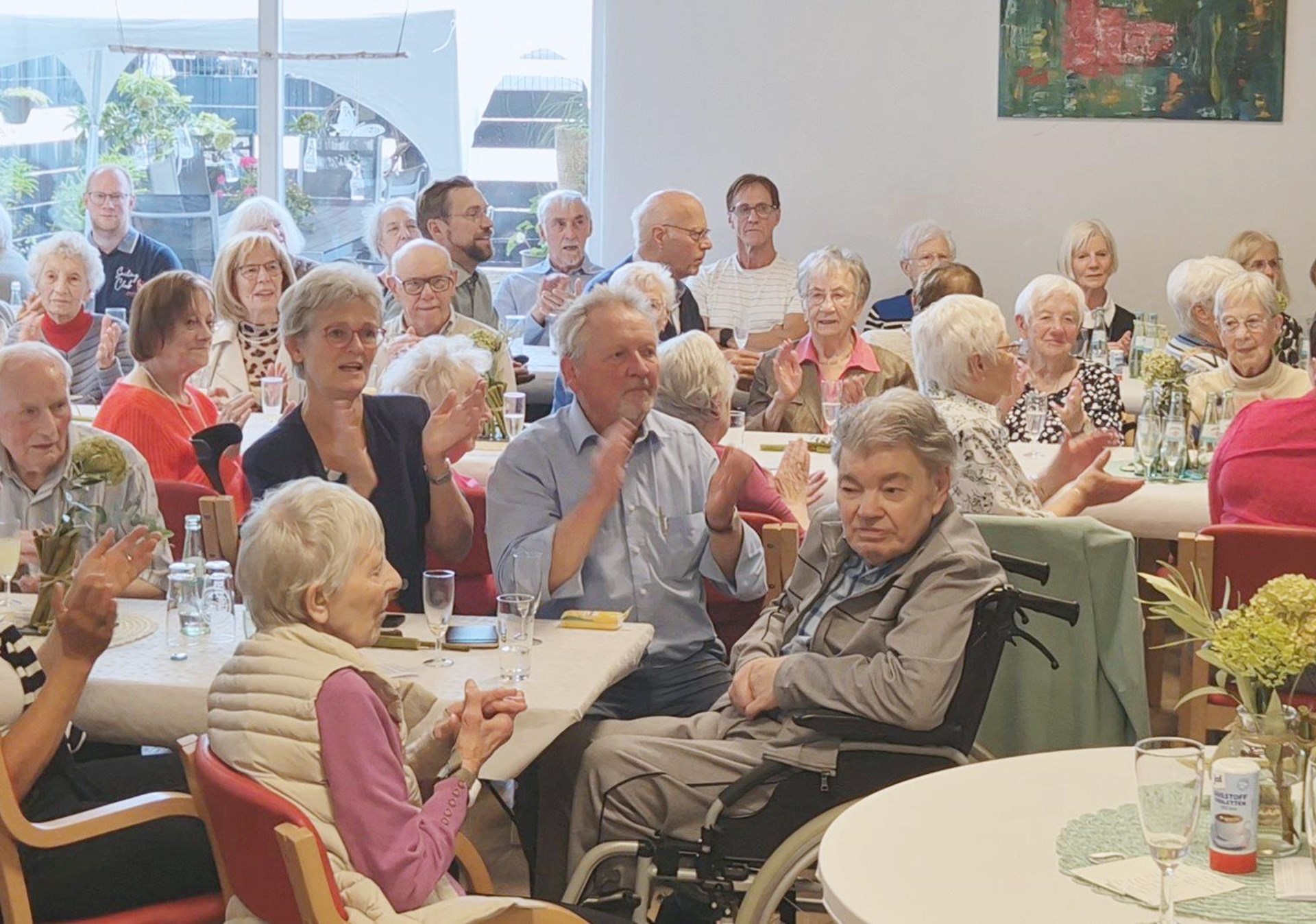 Gäste einer Jubiläumsfeier sitzen gemeinsam an gedeckten Tischen und applaudieren. Unter den zahlreichen Gästen sind ältere Menschen sowie eine Person im Rollstuhl zu sehen.